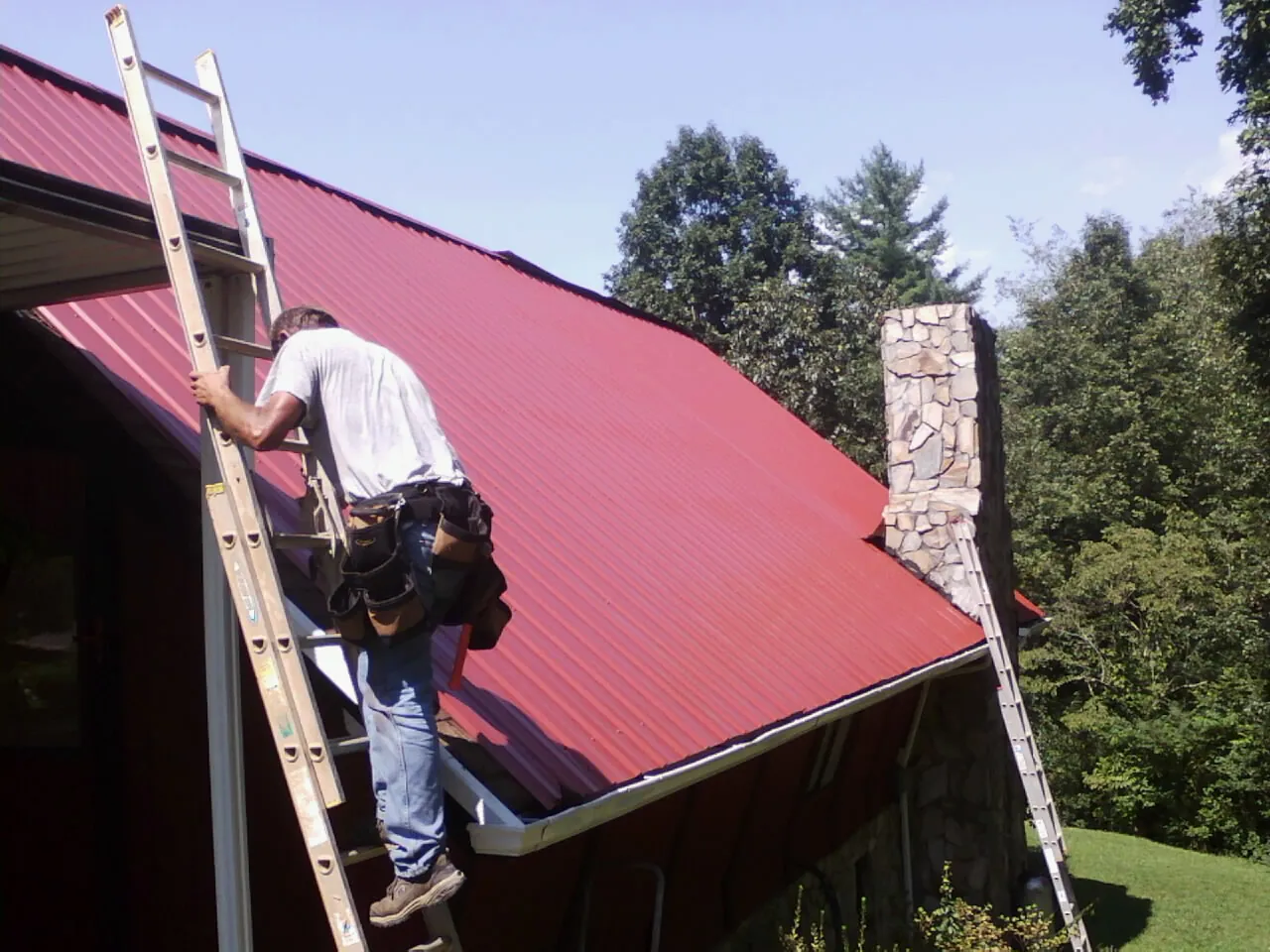 Roofer on a ladder beside a red metal roof and stone chimney — Soffit & Fascia Repair by Roofing Brothers in Shelocta